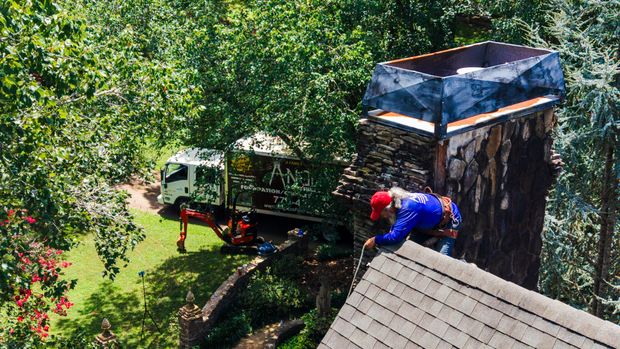 An Anglin's mason working at the top of a tall stone chimney, with the company truck and a mini excavator on the lawn below.