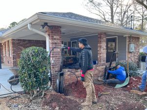 Anglin's crew driving steel piers at the carport column, one at the hydraulic controls while another works the ram against the brick.