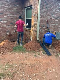 Anglin's crew digging out along the brick wall at the corner of the house, downspout extension pulled aside.