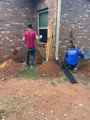 Anglin's crew digging out along the brick wall at the corner of the house, downspout extension pulled aside.