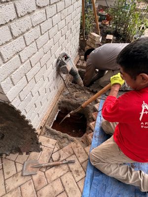 Two crew at the base of the white brick wall, one running a rotary hammer while the other clears the hole, patio pavers lifted and stacked.