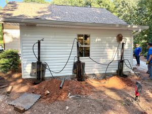 Three hydraulic rams braced against the side of the outbuilding, driving steel piers beside the slab.