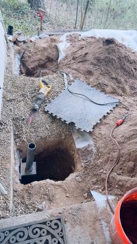 A pier tube standing in its hole with the bracket in place, the wooded slope rising behind the house.