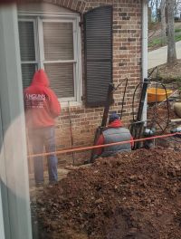 Two crew working the pier at the brick wall below a window, hoses running down into the excavation and the spoil piled in front.