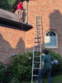 One crew member on the lower roof working the brick wall above it, another steadying the ladder from the flower bed below.