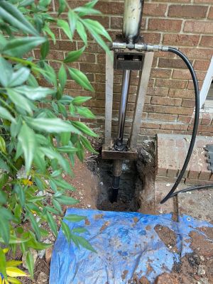 A hydraulic ram driving a steel pier beside the brick foundation, the excavation open beneath it.