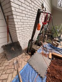 The hydraulic ram set against the wall in the opened hole, hoses and control box alongside, mats and a tarp protecting the pavers.