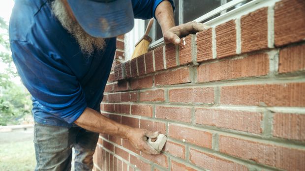 An Anglin's mason working fresh mortar into the joints of a red brick wall beside a window