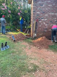 Steel pier tubes and base plates staged on the lawn while the crew opens footing holes along the wall.