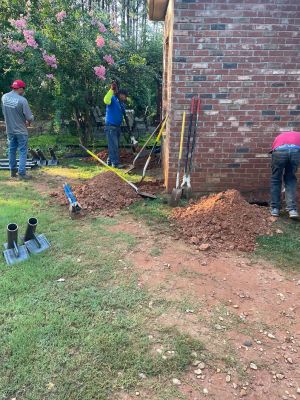 Steel pier tubes and base plates staged on the lawn while the crew opens footing holes along the wall.
