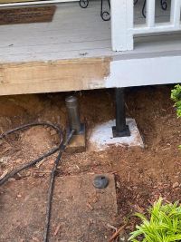 A steel pier set on a fresh concrete footing under the sinking front porch, hydraulic ram on a timber block beside it.