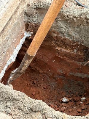 Down into the open pier hole beside the footing, a digging bar working the red clay.