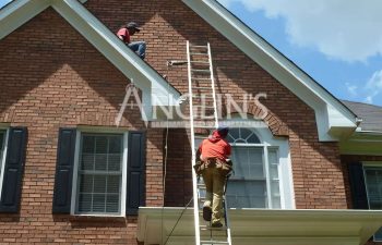 Two workers on ladders are inspecting a two-story brick house with a gabled roof under a blue sky.