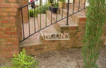 Brick steps with a metal handrail lead up to a porch. Potted plants are on the porch and steps. Shrubs and mulch are in the foreground.